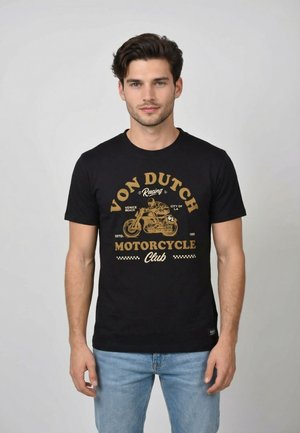 Hombre con camiseta negra con una motocicleta dorada vintage y el texto "Von Dutch Racing Motorcycle Club" de pie frente a un fondo liso.