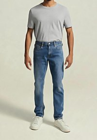 Homme portant un t-shirt gris clair à manches courtes, un jean bleu et des baskets blanches, debout devant un fond clair uni.