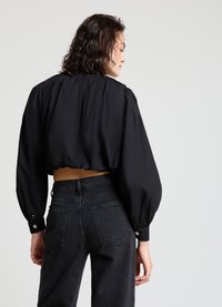 Schwarze, kurz geschnittene Bluse mit Puffärmeln und knöpfbaren Manschetten, kombiniert mit hoch taillierten schwarzen Jeans. Glatte Textur und maßgeschneiderte Passform.