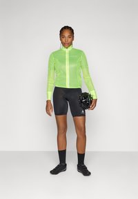 Giacca da ciclismo leggera e traslucida di colore verde, con cerniera full zip, abbinata a pantaloni corti neri imbottiti e scarpe da ciclismo nere e slanciate.