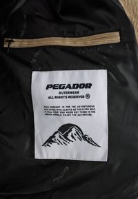 Juodas audinys su šviesiai rudos spalvos užtrauktuku. Aiškiai matomas etiketė, kuri turi kalnų grafiką, prekės ženklo logotipą "PegaDOR" ir priežiūros instrukcijas juodu tekstu.