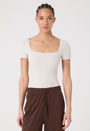 Giovane donna con capelli intrecciati che indossa una maglietta bianca a maniche corte con scollo quadrato e pantaloni marroni con coulisse su uno sfondo semplice.