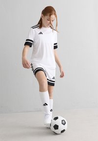 Joven con un uniforme de fútbol blanco y negro controlando un balón de fútbol blanco y negro en interiores, frente a una pared blanca.