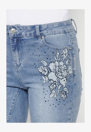 Šviesiai mėlynų džinsų su gėlių siuvinėjimu mėlynomis ir juodomis spalvomis, papuoštų juodais studais prie priekinio kišenės.