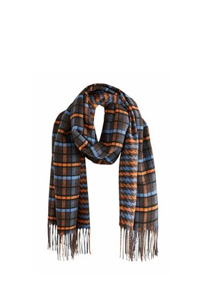 Young woman wearing a dark coat, white shirt, blue jeans, and a long blue and orange striped scarf stands against a brick wall background.