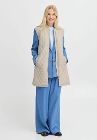 Blonde Frau steht lächelnd da, die Hände in den Taschen, trägt eine beige Steppweste über einem hellblauen Blazer und weit geschnittenen Hosen mit schwarzen Schuhen.