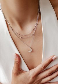 Collana a strati in argento con pendente a mezzaluna, caratterizzata da catene sottili e piccoli accenti sferici, su uno sfondo di tessuto chiaro.
