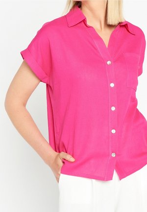 Kvinde iført en lyserød kortærmet skjorte med knapper, krave og lomme, hånd i lommen på hvide bukser.