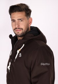 Jachetă capucioasă maro cu închidere cu fermoar, având șnururi albe, logo pe piept și text pe mânecă. Textură netedă, design casual.