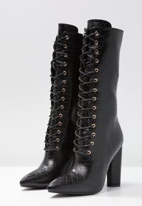 Bottes noires montantes jusqu'au genou en cuir embossé façon crocodile, avec un bout pointu, un talon bloc haut et un devant à lacets avec œillets dorés.