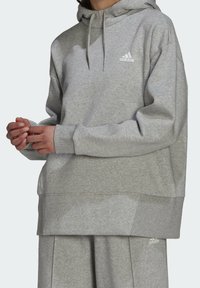 Personne portant un sweat à capuche Adidas gris clair et un pantalon de survêtement assorti avec le logo sur la poitrine et la cuisse, debout les mains jointes.