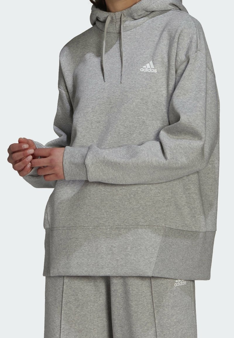 Personne portant un sweat à capuche Adidas gris clair et un pantalon de survêtement assorti avec le logo sur la poitrine et la cuisse, debout les mains jointes.