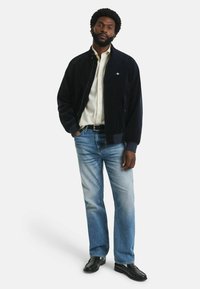 Marineblaue Corduroy-Bomberjacke mit gerippten Bündchen, cremefarbenes Hemd mit Knöpfen, hellblaue Jeans und schwarze Lederschuhe.