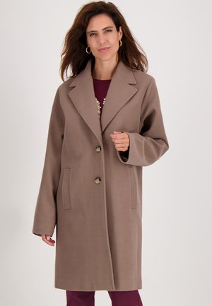 Vrouw in een lange taupe jas met twee knopen over een kastanjekleurige top en broek, met gouden creolen en loshangend bruin haar.