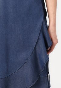 Robe en denim avec un design en couches, un ourlet ondulé, de couleur bleu foncé et une texture lisse. Une main est visible près du bord du tissu.