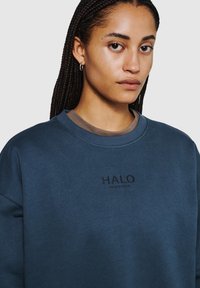 Ciemnozielona bluza z okrągłym dekoltem, z wyhaftowanym czarnym napisem "HALO". Materiał wydaje się miękki, a krój jest luźny.
