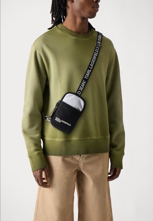 Schwarze Umhängetasche mit glatter Oberfläche und einem weißen Oberelement, ausgestattet mit einem Reißverschluss und einem bedruckten Logo, getragen über einem olivgrünen Sweatshirt.