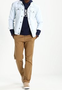 Giacca di jeans azzurro chiaro, felpa blu navy, pantaloni chinos color cammello e sneaker beige. La giacca presenta una chiusura con bottoni e due tasche sul petto.