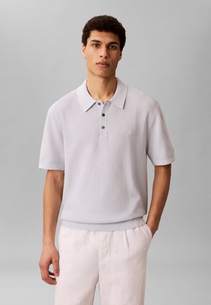 Jonge man met kort krullend haar, die een lichtgrijs gestructureerd poloshirt en witte broek draagt, staand tegen een effen grijze achtergrond.