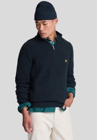 Suéter de canalé azul oscuro con un cuello de media cremallera, combinado con una camisa de botones color teal. Presenta un pequeño logotipo amarillo en el pecho.