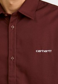 Μπορντό πουκάμισο με σχέδιο κουμπιών, κοντό γιακά και λογότυπο Carhartt σε λευκό χρώμα στο αριστερό στήθος. Φτιαγμένο από λεία, υφασμένη ύφανση.