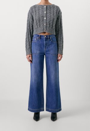 Person trägt eine graue, kurz geschnittene Strickjacke mit Zopfmuster und Knöpfen vorne, hoch taillierte, weit geschnittene blaue Jeans und schwarze Stiefel mit spitzer Zehenpartie.