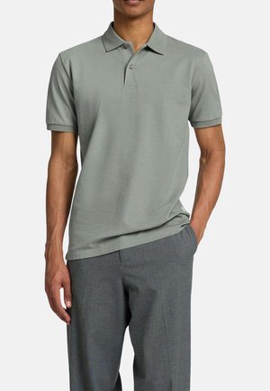 Polo vert clair en tissu doux, avec un col classique, une patte de boutonnage à trois boutons et des manches courtes. Porté avec un pantalon gris.
