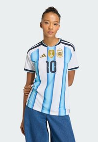 Žena se svázanými vlasy, oblečená v argentinském fotbalovém dresu s číslem 10 a nášivkou FIFA 2023, v kombinaci s modrými džínami, na jednoduchém pozadí.