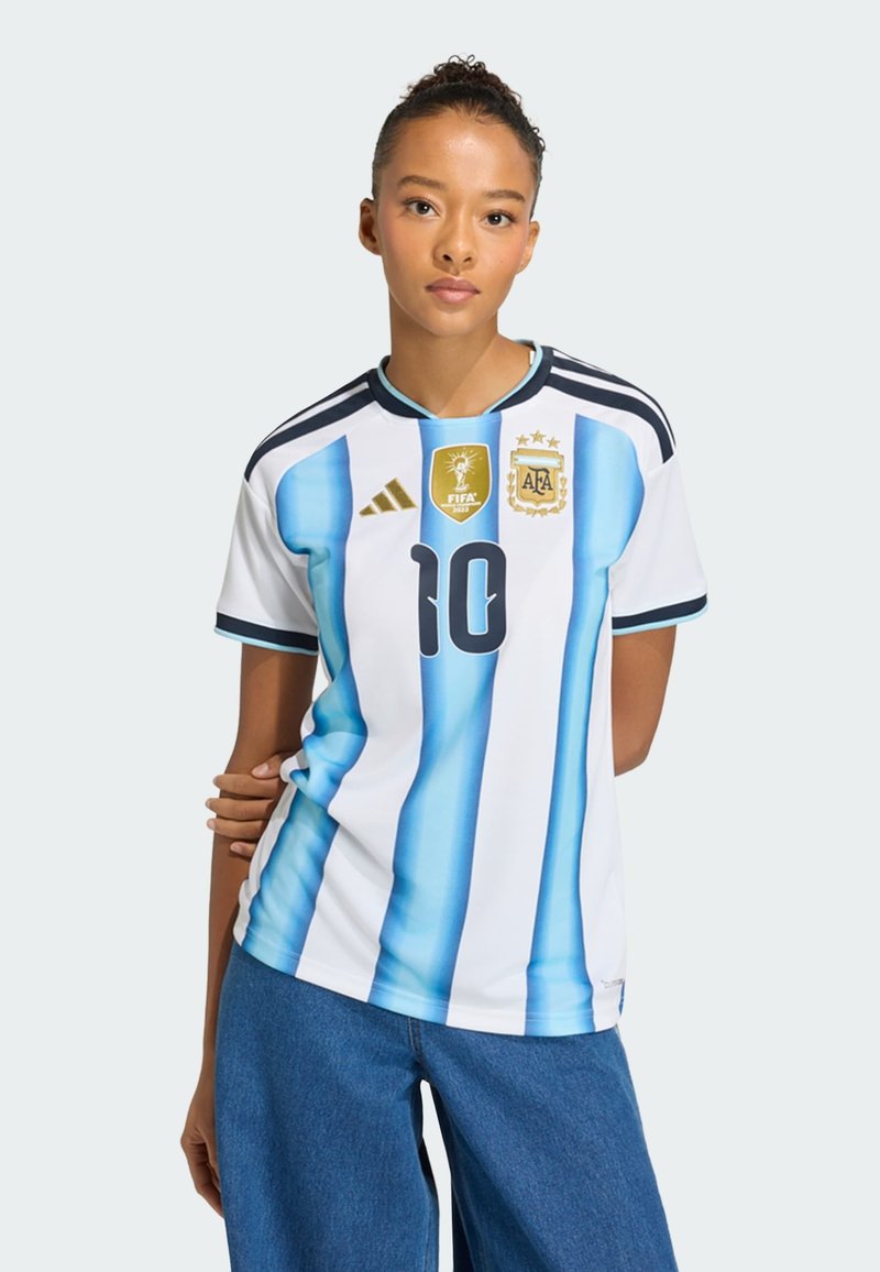 Žena se svázanými vlasy, oblečená v argentinském fotbalovém dresu s číslem 10 a nášivkou FIFA 2023, v kombinaci s modrými džínami, na jednoduchém pozadí.