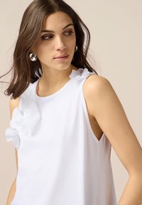 Top bianco senza maniche con fiori in tessuto tridimensionale sulla spalla sinistra. Texture morbida con una vestibilità rilassata e scollatura rotonda.