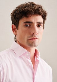 Una camicia button-up rosa chiaro presenta un tessuto testurizzato, colletto standard e bottoni bianchi, indossata da una persona, che mostra un primo piano del torso e del viso.
