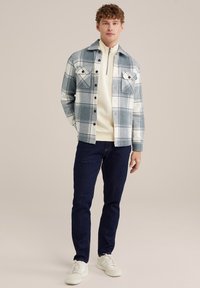 Giacca a quadri in blu chiaro e bianco, con due tasche sul petto e bottoni neri, indossata sopra una maglia con zip crema, abbinata a jeans scuri.