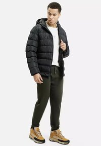 Czarna kurtka puchowa z poziomym pikowaniem, z zamkiem błyskawicznym z przodu i kapturem, noszona na białej koszuli i zielonych dresach, uzupełniona żółtymi butami.