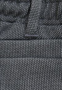 Close-up van grijs en zwart houndstooth-stof met een gestikte lus en een horizontale naaddetail.
