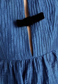 Robe en tissu bleu texturé avec une fente verticale maintenue par un petit nœud noir à l'encolure et une taille froncée.