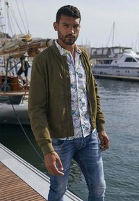 Olijfgroene bomberjack over een witte bloesemshirt, blauwe versleten jeans, staand bij water met boten op de achtergrond.