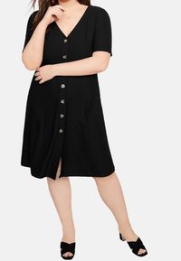 Czarna sukienka z prążkowanego materiału, z krótkimi rękawami, dekoltem w serek i zamknięciem na guziki z przodu. Posiada długość do kolan oraz subtelnie teksturowaną powierzchnię.