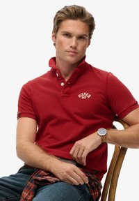 Polo rouge à manches courtes avec un col boutonné et un logo blanc. Le modèle porte une montre marron et un jean en denim, assis sur une chaise en bois.