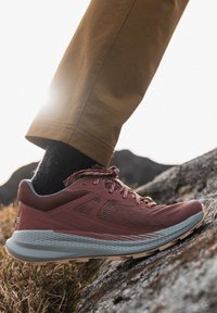 Zapatilla deportiva color marrón con una parte superior de malla transpirable, acentos texturizados, suela gris y cordones marrón claro, que se lleva con pantalones marrones y calcetines negros.