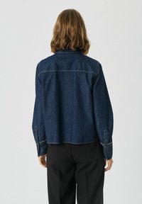 Donkerblauwe spijkerjacket met een cropped ontwerp, met knoopmanchetten en zilveren accenten op de mouwen. Achteraanzicht toont een klassieke kraag en stiksels.