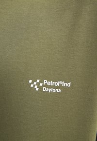 Tela de color verde oliva con una textura suave; presenta la marca en blanco "Petrol Ind" y "Daytona" en la parte inferior derecha.