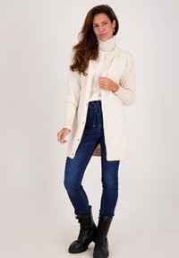 Lange crèmekleurige cardigan gemaakt van zacht materiaal, met knoopsluitingen en geribde mouwen. Draag het over een witte coltrui en combineer het met blauwe jeans en zwarte laarzen.