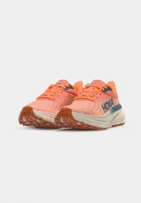 Hoka running shoes in bright coral with a zigzag pattern, featuring orange laces, a cushioned sole, and a contrasting blue accent.