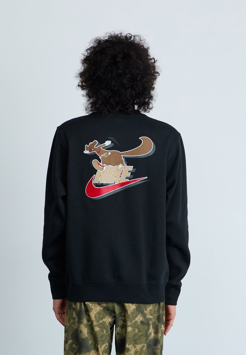 Personne aux cheveux bouclés portant un sweat-shirt noir avec un kangourou de dessin animé courant et un logo rouge Nike dans le dos, associé à un pantalon camouflage vert.