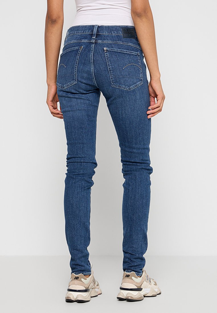 Jean skinny en denim bleu avec une taille haute, dotée de poches arrière et de détails de couture subtils. Associé à des baskets de couleur claire.