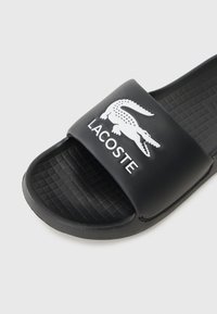 Black slide sandal with a textured sole, featuring a wide strap emblazoned with a white crocodile logo and "LACOSTE" text.
