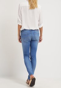 Mujer con cabello largo y rubio que lleva una blusa blanca arremangada, jeans ajustados azul y sandalias negras, caminando de espaldas frente a un fondo liso.