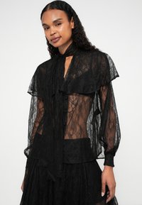 Černá krajková blůza s průsvitnou tkaninou, volánovými detaily, vysokým límcem a dlouhými rukávy. Má knoflíkovou přední část a splývavý střih.