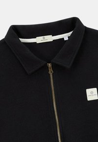 Veste zippée en polaire noire avec un col plat, une fermeture à glissière dorée et une petite étiquette blanche portant "Serge Blanco". Texture douce et design structuré.