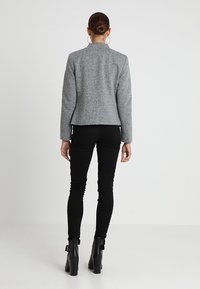 Šedý texturovaný blazer s vypasovaným střihem, má vysoký límec a dlouhé rukávy, doplněný černými skinny džíny a kotníkovými botami.